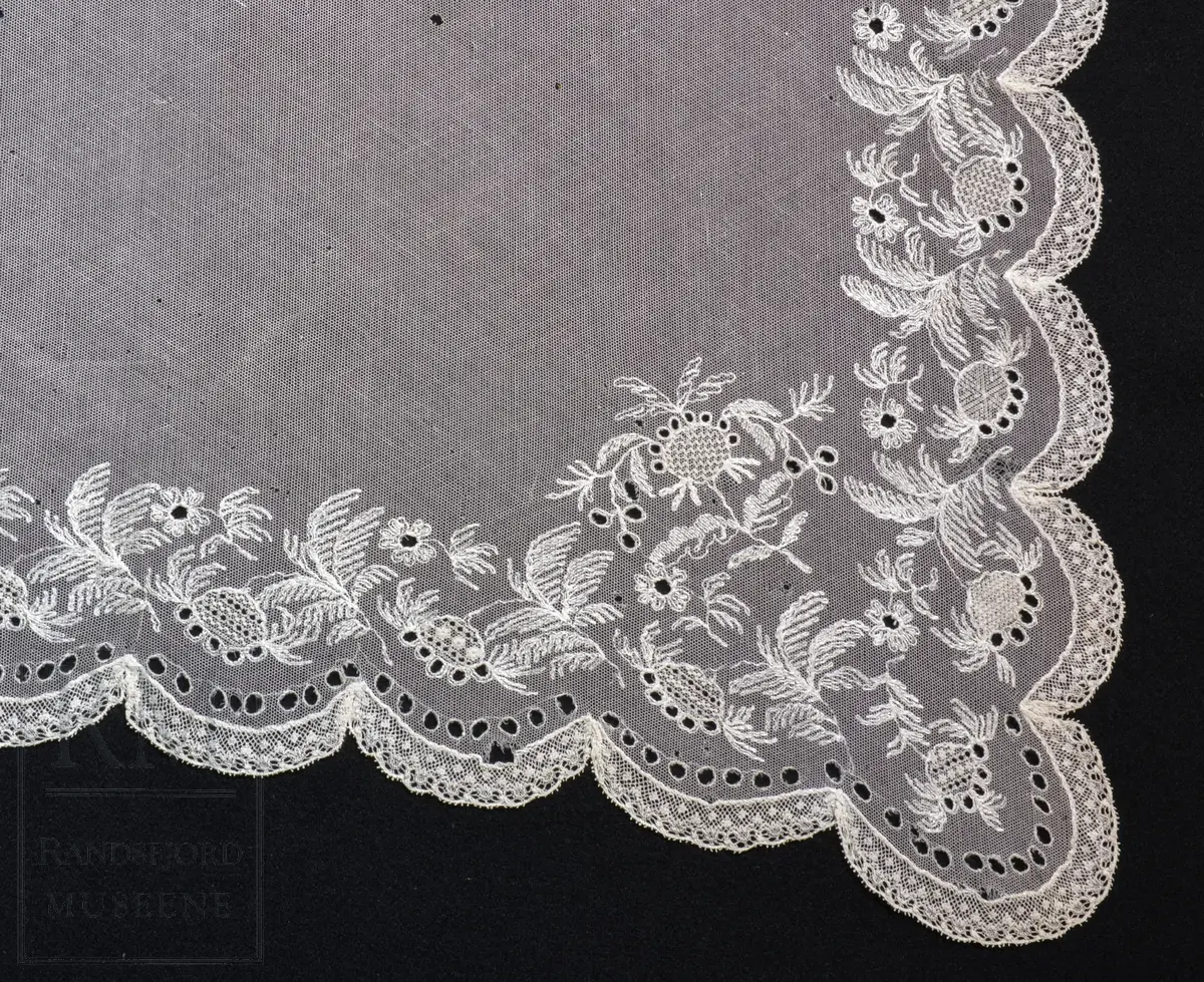 Kvadratisk form, med tungekant. Kantet med kniplingblonde. Tyllbroderi: blomsterranke langs sidene. Større hjørnemotiv i fire hjørner, der et er vesentlig større.  