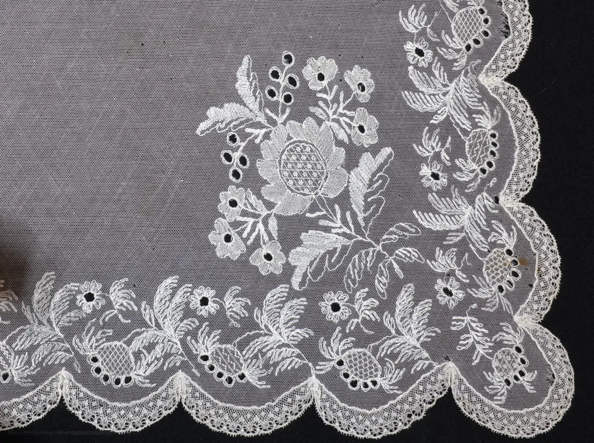 Kvadratisk form, med tungekant. Kantet med kniplingblonde. Tyllbroderi: blomsterranke langs sidene. Større hjørnemotiv i fire hjørner, der et er vesentlig større.  