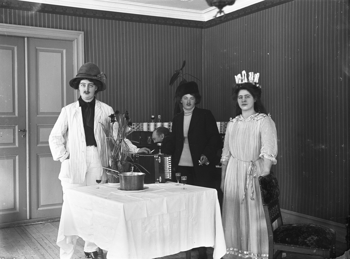 Fest eller en sketch, troligen på Hotell Gästis Varberg. Tre utklädda personer står vid ett bord: en i "tropikhatt", en i kosackliknande mössa och en i luciadräkt med ljuskrona. En man sitter vid pianot i bakgrunden. Kanske är det glögg i grytan med slev på bordet och som hällts upp i glasen. Dragspel och blommor står också på bordet.  Beställare: Ester (Esther) Andersson, som beställt brevkort. En av kvinnorna är troligen beställaren. (Se även bildnr GB2_5406.)