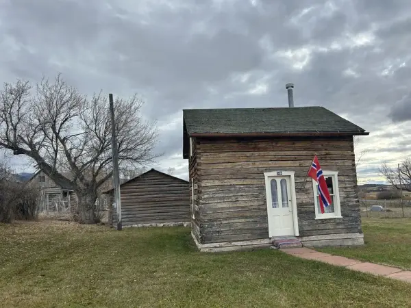 Et lite, laftet hus med norsk flagg ved inngangsdøren. Flere bygninger i bakgrunn. Overskyet vær.