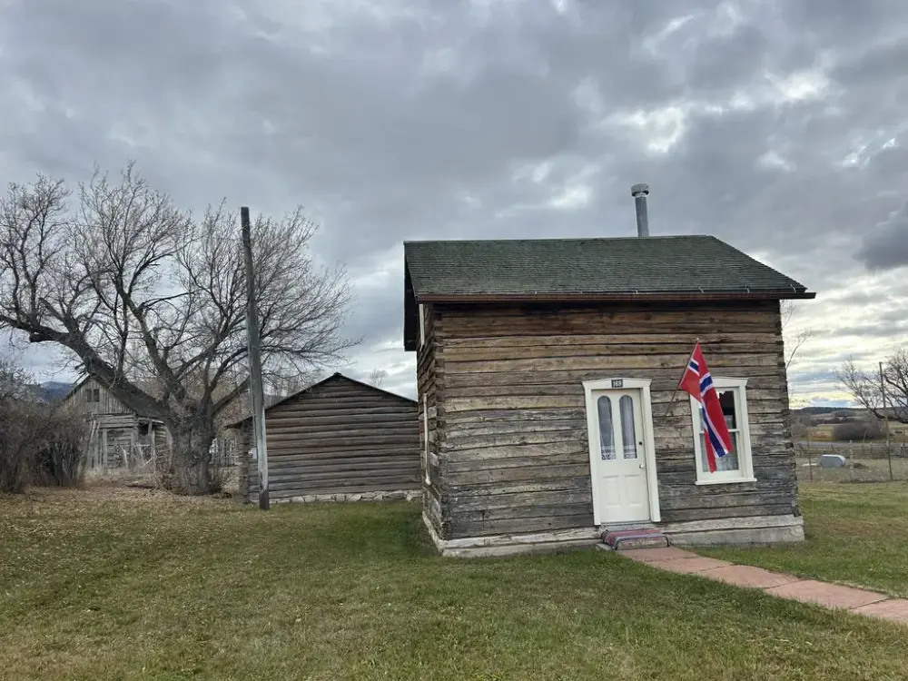Et lite, laftet hus med norsk flagg ved inngangsdøren. Flere bygninger i bakgrunn. Overskyet vær.