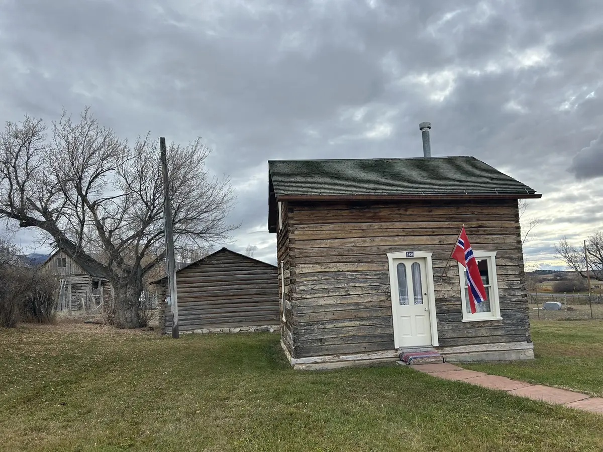 Et lite, laftet hus med norsk flagg ved inngangsdøren. Flere bygninger i bakgrunn. Overskyet vær.