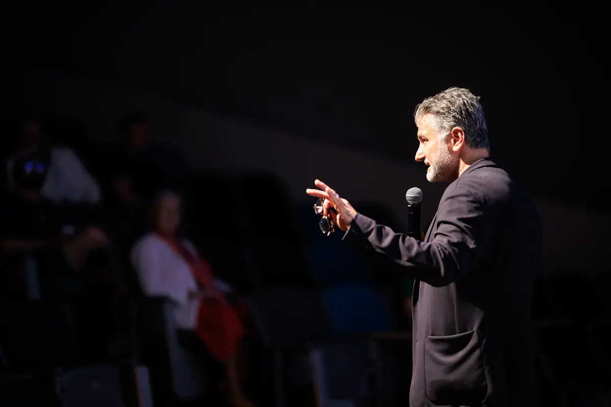 En mann med mikrofon står til høyre. Publikum skimtes til venstre. Mørk bakgrunn.