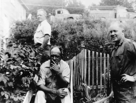 Tre menn i en hage med flere nivåer. Mannen i midten sitter ved en port eller et gjerde. Øverst står to lastebiler.