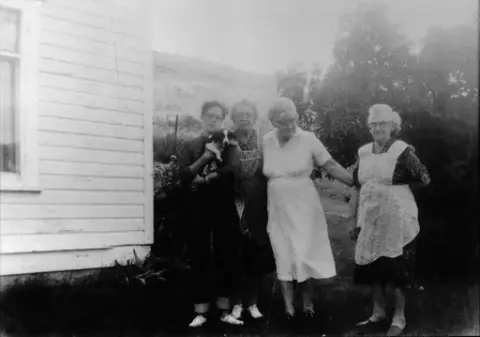 Tre eldre kvinner og en ung mann som holder en valp står i en hage ved en lys husvegg. To av kvinnene har et forkle over kjolen sin. Fjellside i bakgrunn.