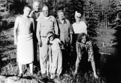 Syv mennesker i skogen, både barn og voksne. Ungdommen til venstre har et håndkle viklet rundt underkroppen. Det miste barnet har krykker og skjermer øynene for solen. Personen bak til høyre har solbriller.