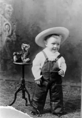 Portrett av en liten gutt med snekkerbukser og en stor hatt. Han står ved siden av en rund krakk som en bamse sitter på.