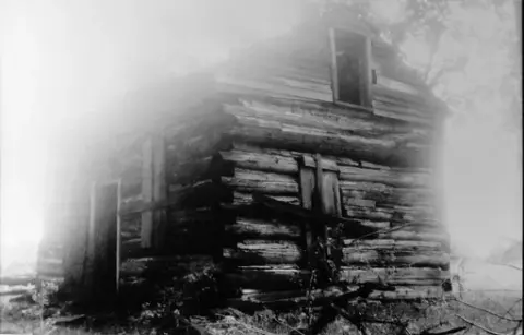 Et lite, laftet hus i svært dårlig forfatning. Vinduene nede er spikret for.