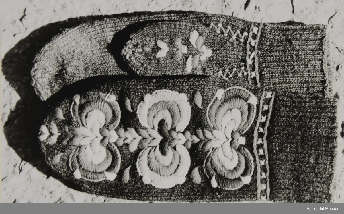 Votter strikka av raudbrunt ullgarn. Stilisert blomstremotiv på framsida, i tjukksaum med flanillsgarn i 3 raudfargar, raudorange, gult, 2 grønfargar og kvitt.