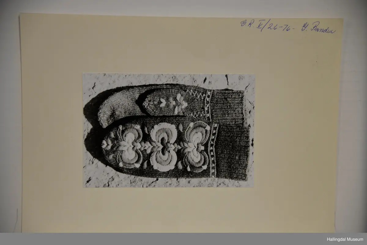 Votter strikka av raudbrunt ullgarn. Stilisert blomstremotiv på framsida, i tjukksaum med flanillsgarn i 3 raudfargar, raudorange, gult, 2 grønfargar og kvitt.