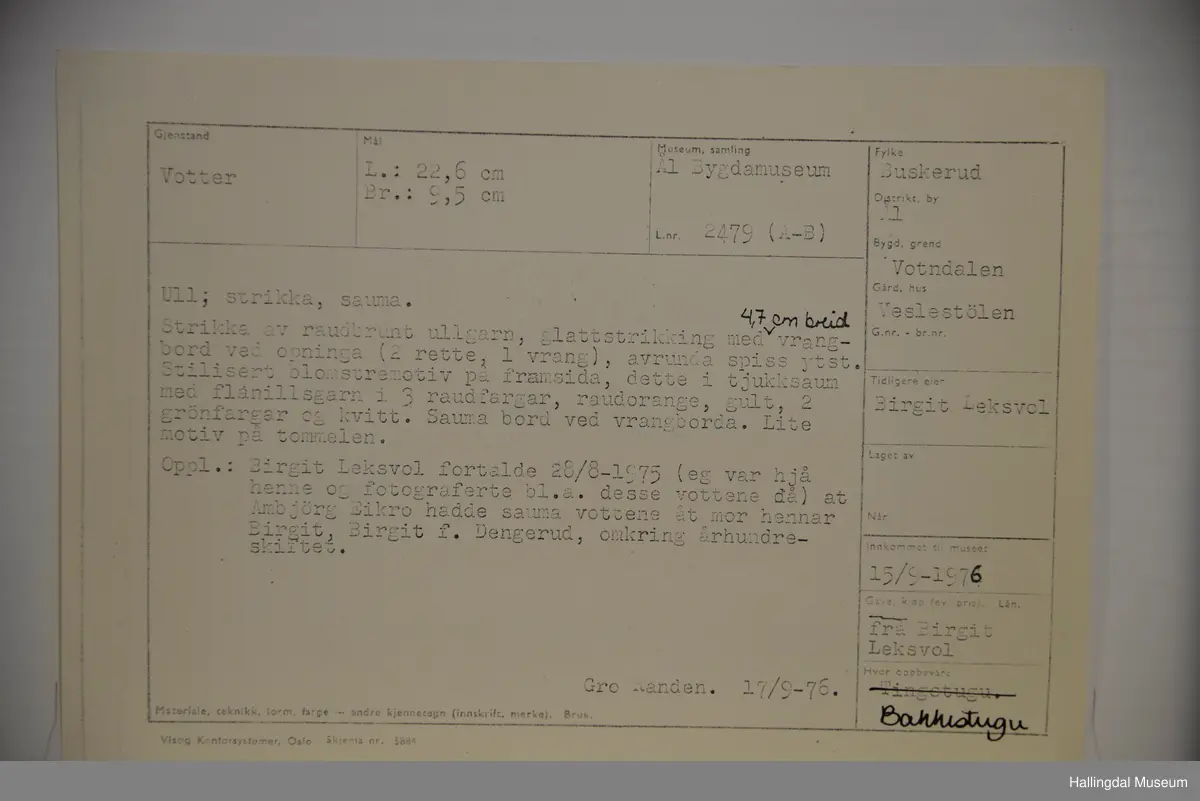 Votter strikka av raudbrunt ullgarn. Stilisert blomstremotiv på framsida, i tjukksaum med flanillsgarn i 3 raudfargar, raudorange, gult, 2 grønfargar og kvitt.
