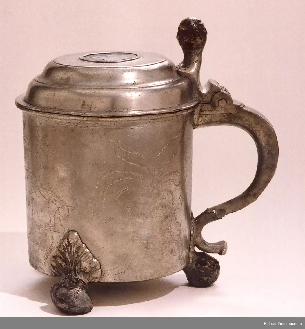 KLM 5411. Dryckeskanna av tenn. Cylindrisk försedd med högt lock med knapp samt tre runda fötter. På yttersidorna bladornament och mittfram en sköld med 2 korslagda C, två lejon med sköldhållare och däröver en kunglig krona. På utsidan av locket en avgjutning av jubelfestmedaljer 17 mars 1721 (H.1,33). Utanpå i botten tre kronor en tenngjutarstämpel, namnet Westerwik samt W (1779).