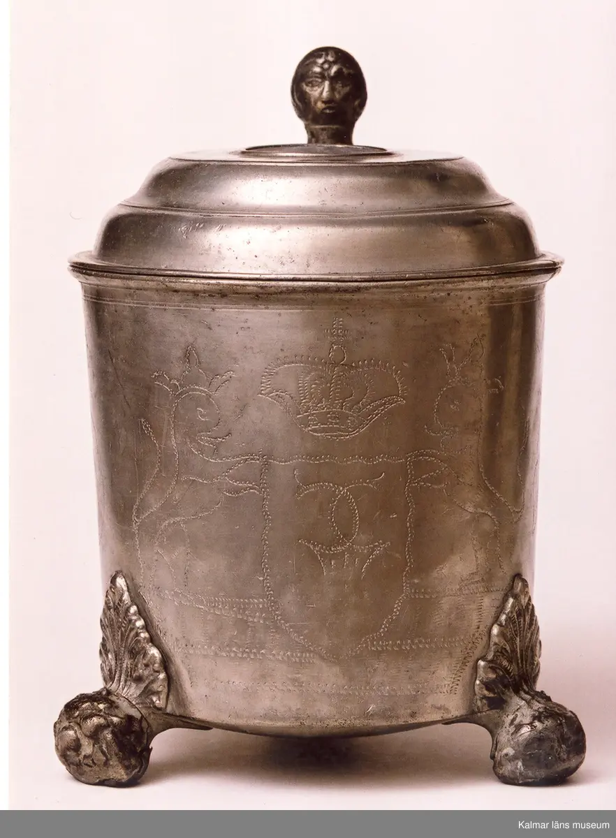KLM 5411. Dryckeskanna av tenn. Cylindrisk försedd med högt lock med knapp samt tre runda fötter. På yttersidorna bladornament och mittfram en sköld med 2 korslagda C, två lejon med sköldhållare och däröver en kunglig krona. På utsidan av locket en avgjutning av jubelfestmedaljer 17 mars 1721 (H.1,33). Utanpå i botten tre kronor en tenngjutarstämpel, namnet Westerwik samt W (1779).