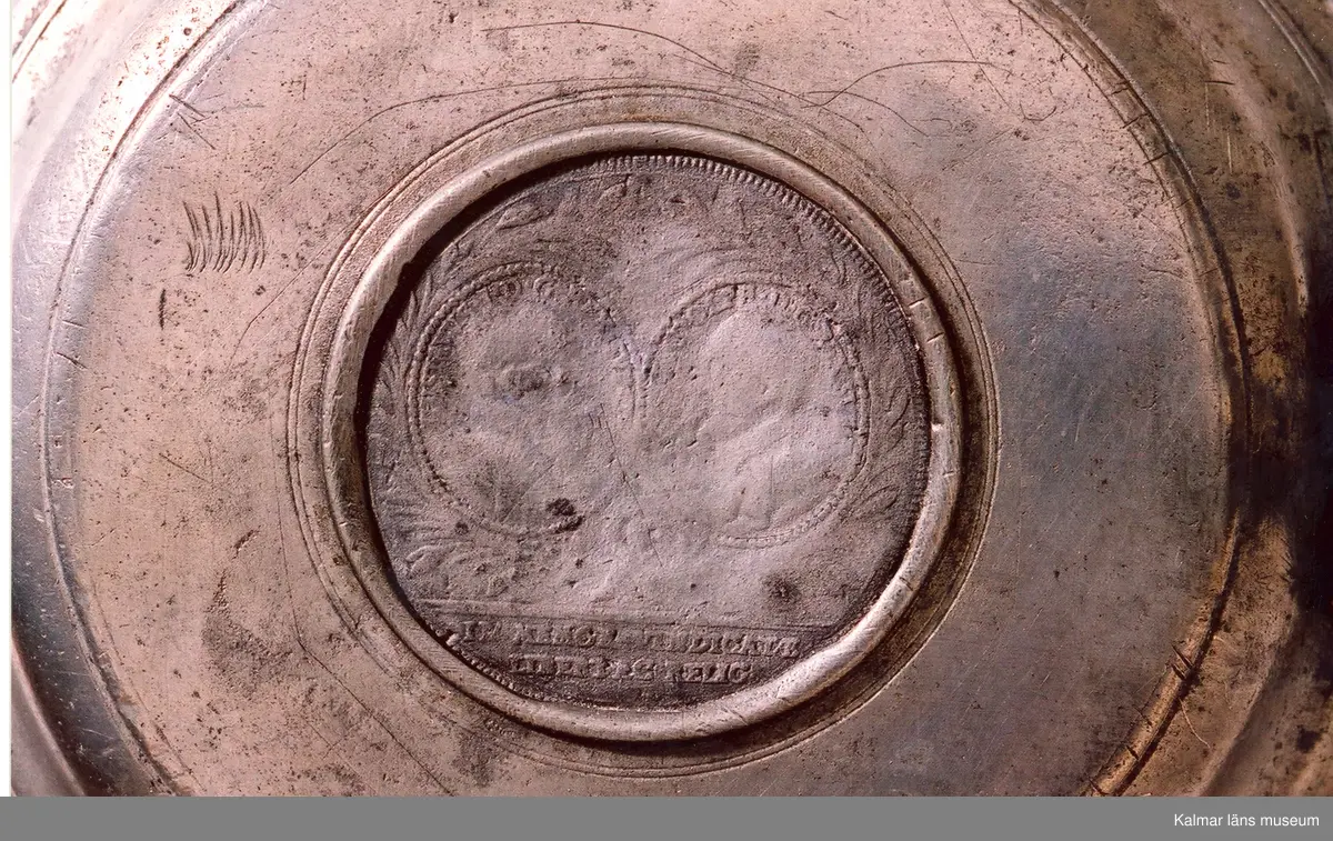 KLM 5411. Dryckeskanna av tenn. Cylindrisk försedd med högt lock med knapp samt tre runda fötter. På yttersidorna bladornament och mittfram en sköld med 2 korslagda C, två lejon med sköldhållare och däröver en kunglig krona. På utsidan av locket en avgjutning av jubelfestmedaljer 17 mars 1721 (H.1,33). Utanpå i botten tre kronor en tenngjutarstämpel, namnet Westerwik samt W (1779).