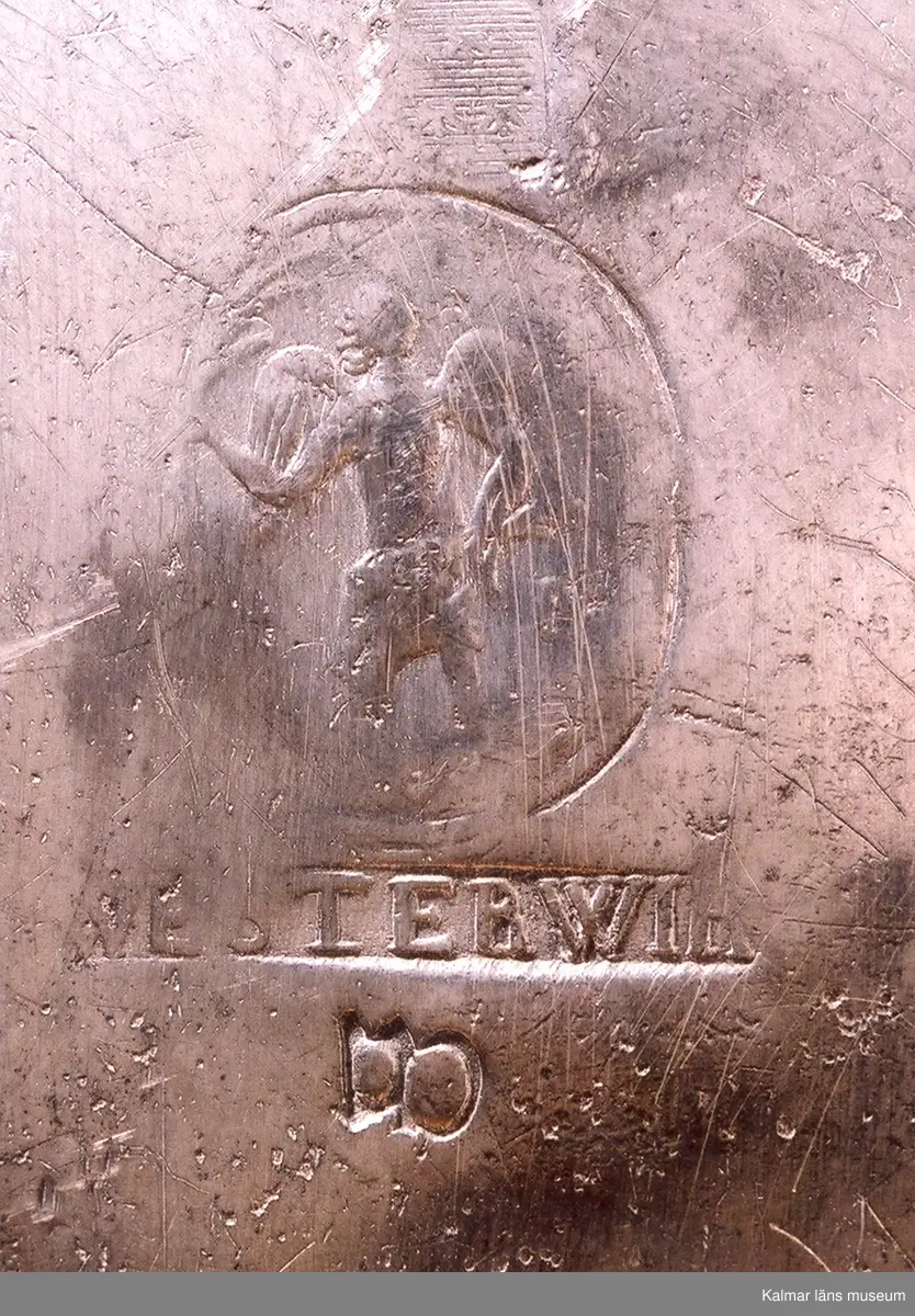 KLM 5411. Dryckeskanna av tenn. Cylindrisk försedd med högt lock med knapp samt tre runda fötter. På yttersidorna bladornament och mittfram en sköld med 2 korslagda C, två lejon med sköldhållare och däröver en kunglig krona. På utsidan av locket en avgjutning av jubelfestmedaljer 17 mars 1721 (H.1,33). Utanpå i botten tre kronor en tenngjutarstämpel, namnet Westerwik samt W (1779).