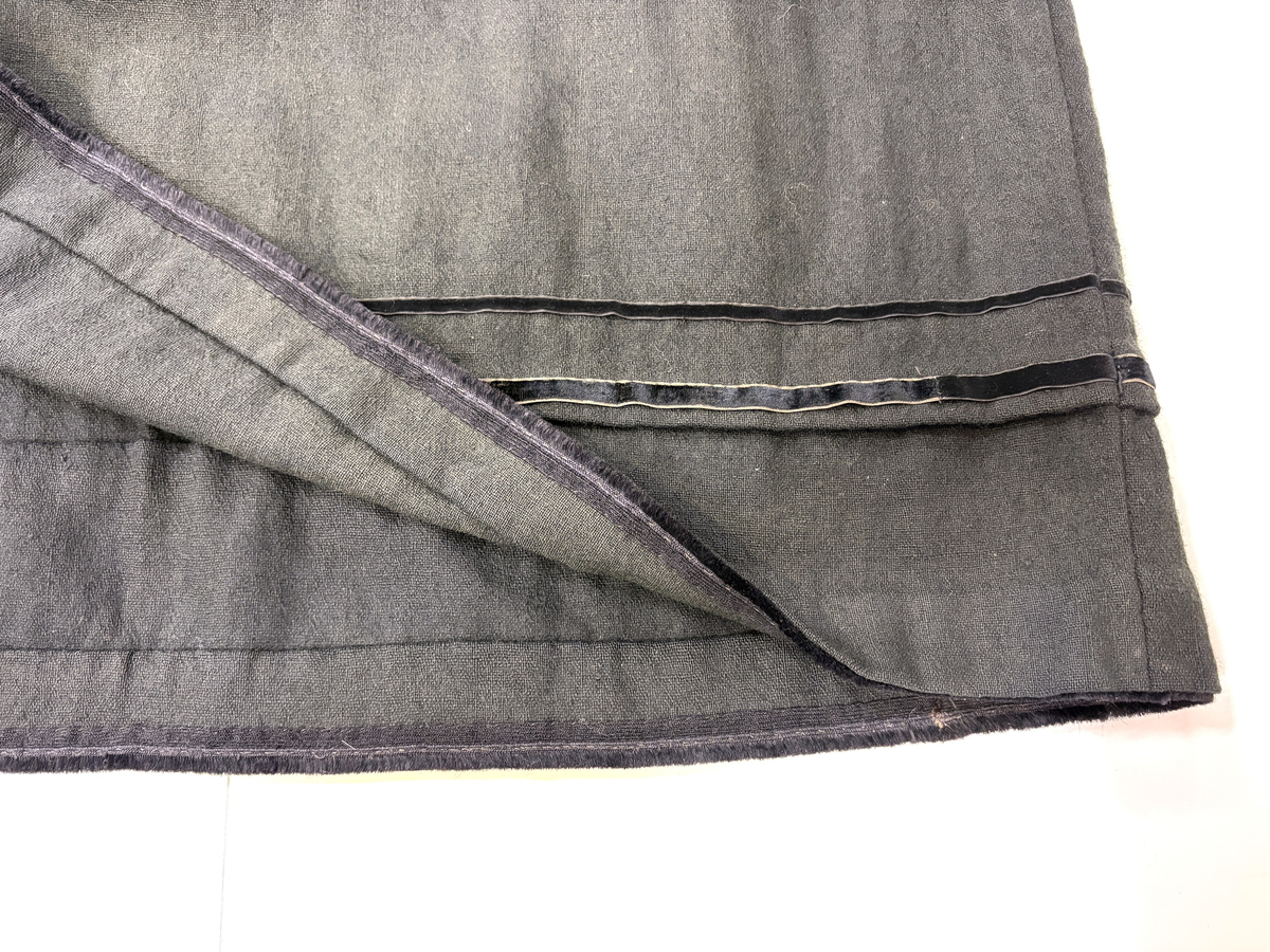 Stakk i samme stoff som liv SM.008024.01; svart ull i hjemmevevd toskaft. Stakken er i god stand med litt slitasje på et pyntebånd. Sydd sammen med maskin med brun bomullstråd av fem vevhøyder med jarekanter. Stakken har et 24 cm bredt glatt stykke midt på foran og foldelagt på begge sider. Åpningen er i venstre side og er 26, 5 cm lang. Stakken har seks folder foldelagt mot høyre og seks folder foldelagt mot venstre, 4-4,5 cm brede. Midt på bak er det et 7,5 cm bredt rynket felt med en halv fold over på begge sider. Foldene er tråklet, sydd på med attersting og så kastet med faldesting med brun bomullstråd. Linningen er i samme stoff som kjolen og 3 cm bred, sydd med faldesting med brun bomullstråd og deretter med prikkesting midt på linningen med samme tråd. Lukkes med to metallhekter. Stakken har en 3 cm bred hengefald 10 cm fra nedkanten, sydd med faldesting med brun bomullstråd. Pyntet nede med to fløyelsbånd som er 0,7 og 1 cm brede, sydd på for hånd i øverste kant av båndet med prikkesting med svart bomullstråd. 0,5 cm bred kostekant i fløyel sydd på med atter- og faldesting for hånd med attersting med svart bomullstråd. Stakken har en 24 cm lang og 17 cm bred lomme på høyre side i samme stoff som fôret på livet, sydd med attersting med svart bomullstråd og festet til stakken med maskin med brun bomullstråd. Stakken har en metallhempe midt på bak på linningen for å henge på livet, og stakken skal brukes ned i livet.