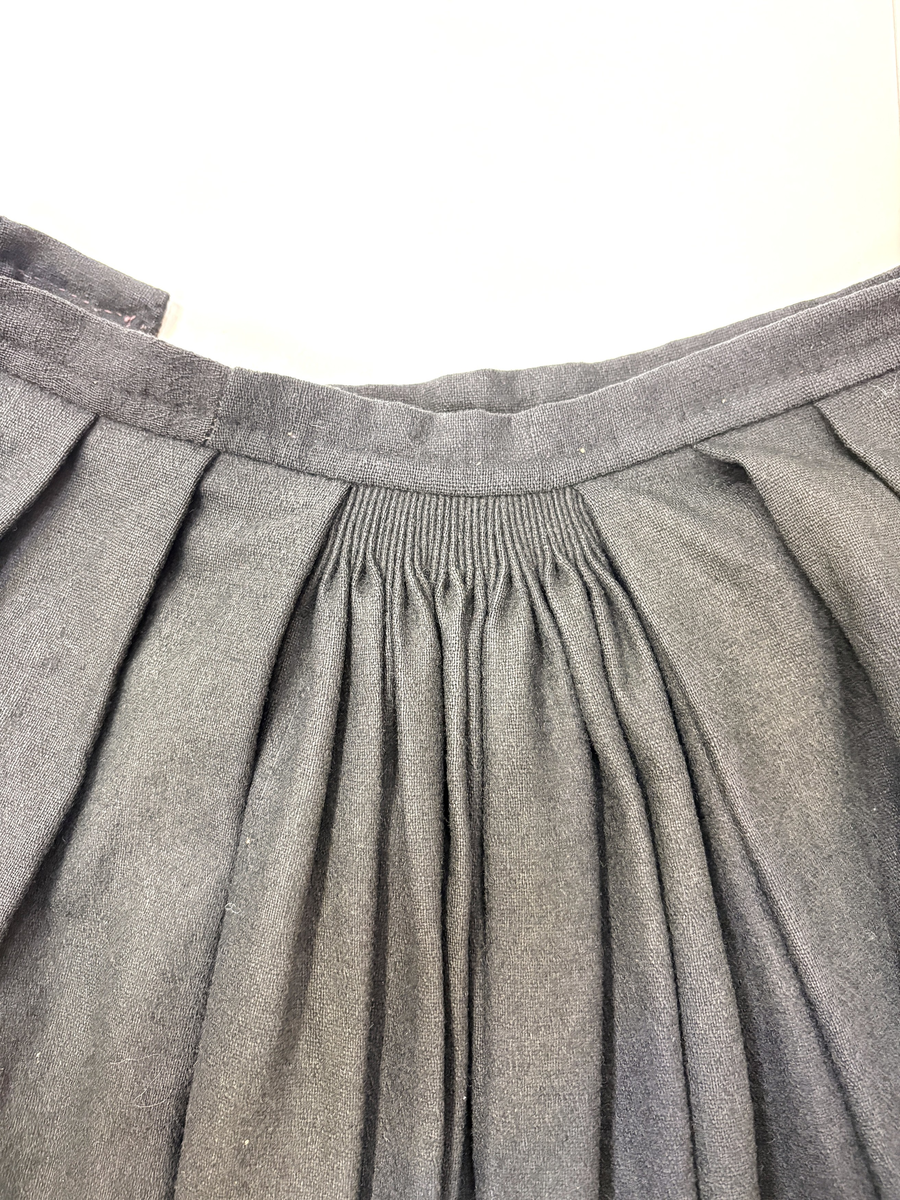 Stakk i samme stoff som liv SM.008024.01; svart ull i hjemmevevd toskaft. Stakken er i god stand med litt slitasje på et pyntebånd. Sydd sammen med maskin med brun bomullstråd av fem vevhøyder med jarekanter. Stakken har et 24 cm bredt glatt stykke midt på foran og foldelagt på begge sider. Åpningen er i venstre side og er 26, 5 cm lang. Stakken har seks folder foldelagt mot høyre og seks folder foldelagt mot venstre, 4-4,5 cm brede. Midt på bak er det et 7,5 cm bredt rynket felt med en halv fold over på begge sider. Foldene er tråklet, sydd på med attersting og så kastet med faldesting med brun bomullstråd. Linningen er i samme stoff som kjolen og 3 cm bred, sydd med faldesting med brun bomullstråd og deretter med prikkesting midt på linningen med samme tråd. Lukkes med to metallhekter. Stakken har en 3 cm bred hengefald 10 cm fra nedkanten, sydd med faldesting med brun bomullstråd. Pyntet nede med to fløyelsbånd som er 0,7 og 1 cm brede, sydd på for hånd i øverste kant av båndet med prikkesting med svart bomullstråd. 0,5 cm bred kostekant i fløyel sydd på med atter- og faldesting for hånd med attersting med svart bomullstråd. Stakken har en 24 cm lang og 17 cm bred lomme på høyre side i samme stoff som fôret på livet, sydd med attersting med svart bomullstråd og festet til stakken med maskin med brun bomullstråd. Stakken har en metallhempe midt på bak på linningen for å henge på livet, og stakken skal brukes ned i livet.