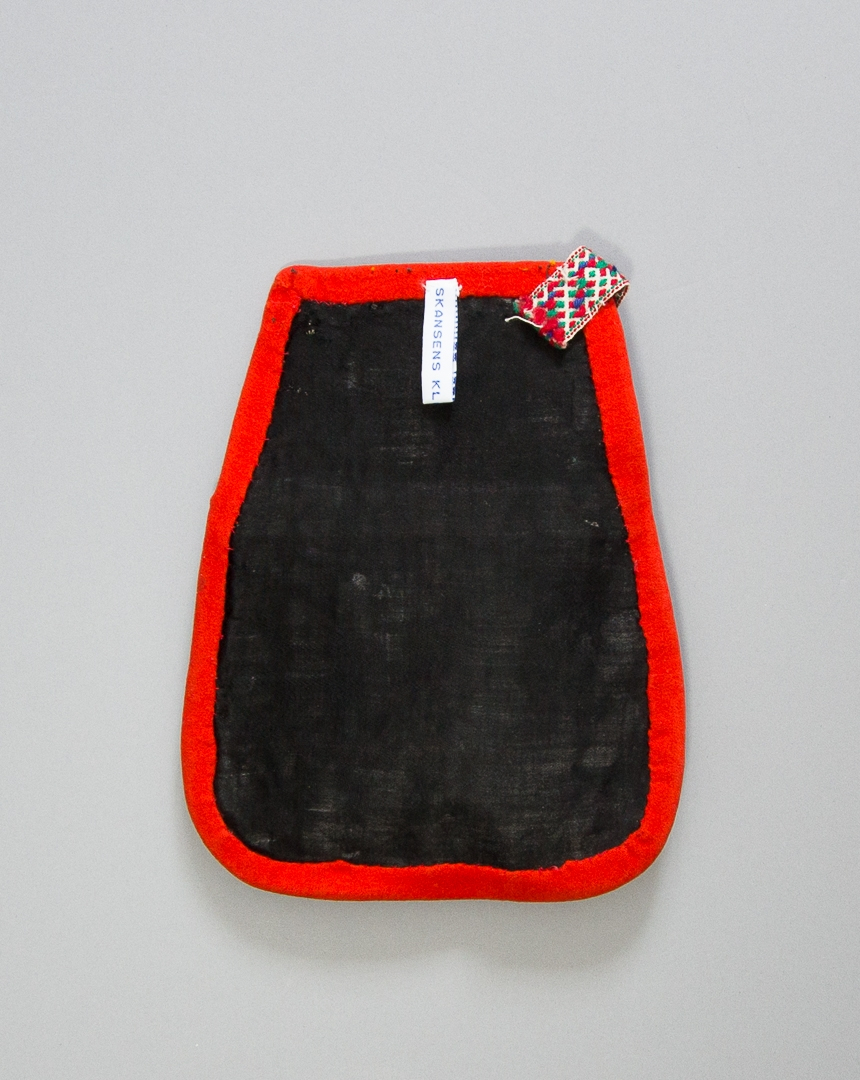 Kjolsäck till dräkt för kvinna från Gagnefs socken, Dalarna. Modell med avskuret framstycke. Tillverkad av svart ylletyg vävt i kypert, med applikationer av kläde i rött, grönt och orange, fastsydda med läggsöm. Centralt placerat hjärtmotiv med slingor och bladmotiv runtom. Broderier utförda med glansigt bomullsgarn i många färger, sticksöm, flätsöm, plattsöm, även på överstycket. Kantad runtom med remsor av rött kläde. Foder av fabriksvävt vitt bomullstyg, lakansväv. Baksida av glansigt svart bomullstyg vävt i satin, med okänt mellanlägg. Midjeband handvävt med plockat mönster av rött, grönt och blått ullgarn på vit botten av lin.