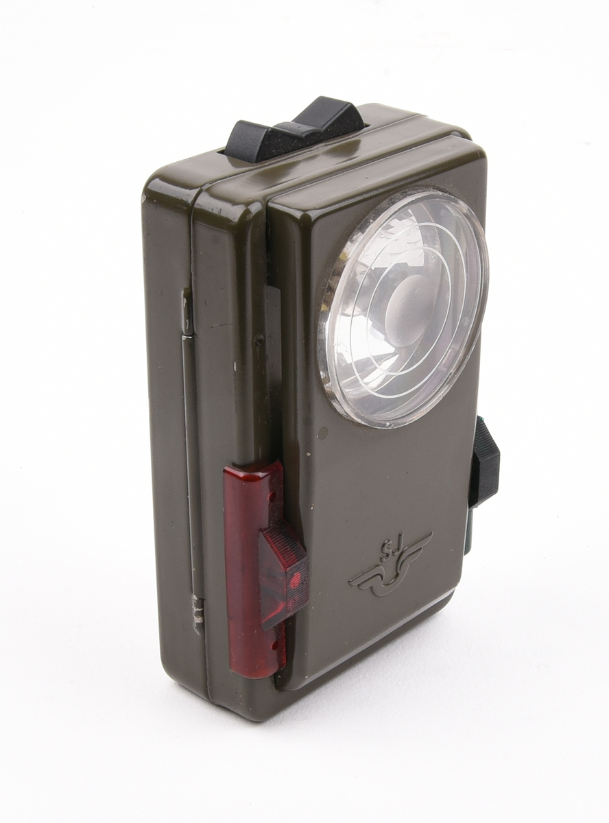 Rektangulär ficklampa av grön metall med signalfunktion. Upptill en svart knapp för att tända och släcka lampan. Framtill en rund, klar lins av plast med en glödlampa innanför. Text "SJ" och SJs logotyp nedanför linsen. Vid sidorna sitter en grön respektive röd knapp för att för skjuta upp ett tunt gröntonat respektive rödtonat plastfilter. Den gröna färgen markerar "Klart för avgång" och den röda färgen "Nödstopp". Baktill tre bruna läderremmar som gör att lampan kan bäras i handen eller fästas vid exempelvis ett bälte.