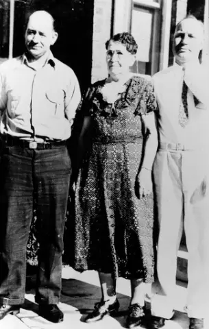 To menn og en kvinne utenfor et hus. Kvinnen har briller og en mønstrete kjole.