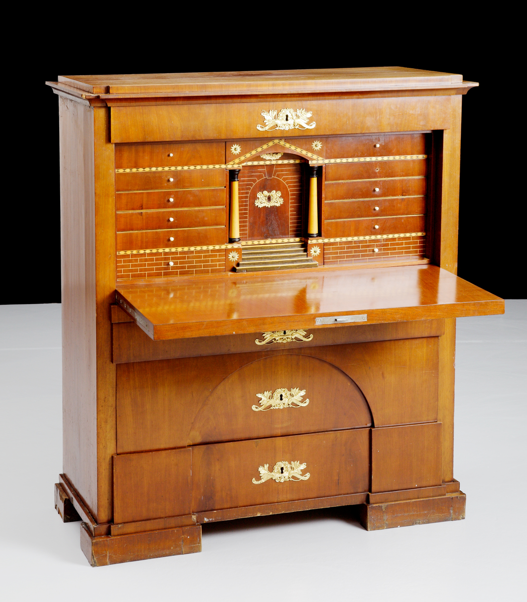 Chiffonje av mahogny. Fanerat och med intarsia på den inre inredningen. Utförd med speciell inredning med flera lönnfack. Lådorna och klaffen har förgyllda beslag. Har varit brukad för förvaring.
Inskriven i huvudkatalog 1943.