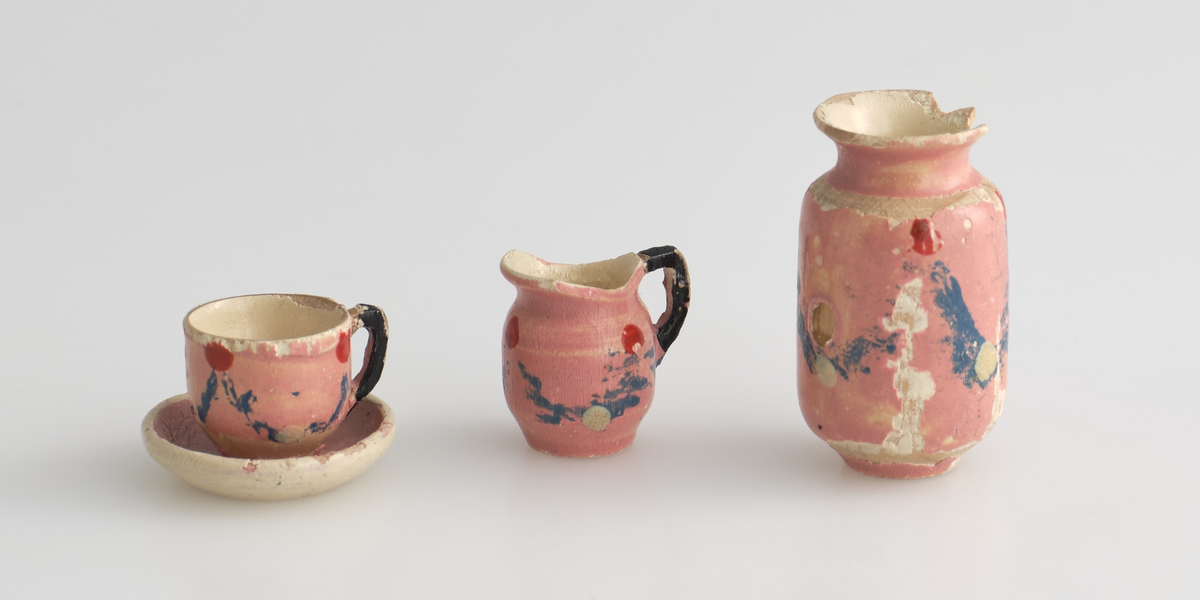 Serveringssett i miniatyr, lagd av tre.
Settet består av en tallerken, en kopp, en mugge og en krukke. Hele settet er malt i samme farger dekorert med likt mønster, utenom tallerkenen som ikke har malt mønster.
Tallerkenen har forhøyet kanter, og er malt rosa på innsiden, med hvite kanter og hvit underside.
Koppen er malt hvit på innsiden, og har en svartmalt hank. Hanken er øreformet og er festet fra kanten øverst til hvor koppen buer seg innover ved foten. Utsiden av koppen er malt rosa, og dekorert med blåe streker som danner trekanter med ujevne linjer rundt koppen. På toppen av trekantene er det malt en rød sirkel.
Krukken er sylinder-formet og snevres inn i mot toppen, før den vies skrått utover rundt åpningen. Krukken er malt hvit på innsiden, og rosa på utsiden, utsiden er også dekorert med samme mønster som koppen. Kanten rundt krukkens åpning er delvis brukket av.
Muggen er sylindret-formet, og snevres inn mot toppen, hvor kanten rundt åpningen forhøyes mot helletuten på den ene siden. På motsatt side av helletuten er det festet en svart-malt hank. Oversiden av hanken er rett, og bues ned mot bunnen av muggen. Muggen er malt og dekorert likt som koppen.