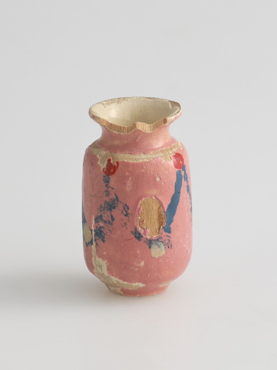 Serveringssett i miniatyr, lagd av tre.
Settet består av en tallerken, en kopp, en mugge og en krukke. Hele settet er malt i samme farger dekorert med likt mønster, utenom tallerkenen som ikke har malt mønster.
Tallerkenen har forhøyet kanter, og er malt rosa på innsiden, med hvite kanter og hvit underside.
Koppen er malt hvit på innsiden, og har en svartmalt hank. Hanken er øreformet og er festet fra kanten øverst til hvor koppen buer seg innover ved foten. Utsiden av koppen er malt rosa, og dekorert med blåe streker som danner trekanter med ujevne linjer rundt koppen. På toppen av trekantene er det malt en rød sirkel.
Krukken er sylinder-formet og snevres inn i mot toppen, før den vies skrått utover rundt åpningen. Krukken er malt hvit på innsiden, og rosa på utsiden, utsiden er også dekorert med samme mønster som koppen. Kanten rundt krukkens åpning er delvis brukket av.
Muggen er sylindret-formet, og snevres inn mot toppen, hvor kanten rundt åpningen forhøyes mot helletuten på den ene siden. På motsatt side av helletuten er det festet en svart-malt hank. Oversiden av hanken er rett, og bues ned mot bunnen av muggen. Muggen er malt og dekorert likt som koppen.