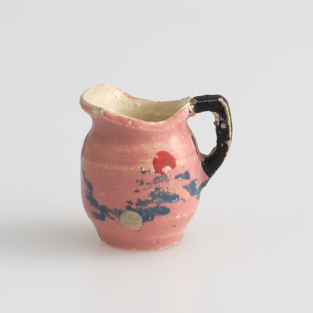 Serveringssett i miniatyr, lagd av tre.
Settet består av en tallerken, en kopp, en mugge og en krukke. Hele settet er malt i samme farger dekorert med likt mønster, utenom tallerkenen som ikke har malt mønster.
Tallerkenen har forhøyet kanter, og er malt rosa på innsiden, med hvite kanter og hvit underside.
Koppen er malt hvit på innsiden, og har en svartmalt hank. Hanken er øreformet og er festet fra kanten øverst til hvor koppen buer seg innover ved foten. Utsiden av koppen er malt rosa, og dekorert med blåe streker som danner trekanter med ujevne linjer rundt koppen. På toppen av trekantene er det malt en rød sirkel.
Krukken er sylinder-formet og snevres inn i mot toppen, før den vies skrått utover rundt åpningen. Krukken er malt hvit på innsiden, og rosa på utsiden, utsiden er også dekorert med samme mønster som koppen. Kanten rundt krukkens åpning er delvis brukket av.
Muggen er sylindret-formet, og snevres inn mot toppen, hvor kanten rundt åpningen forhøyes mot helletuten på den ene siden. På motsatt side av helletuten er det festet en svart-malt hank. Oversiden av hanken er rett, og bues ned mot bunnen av muggen. Muggen er malt og dekorert likt som koppen.