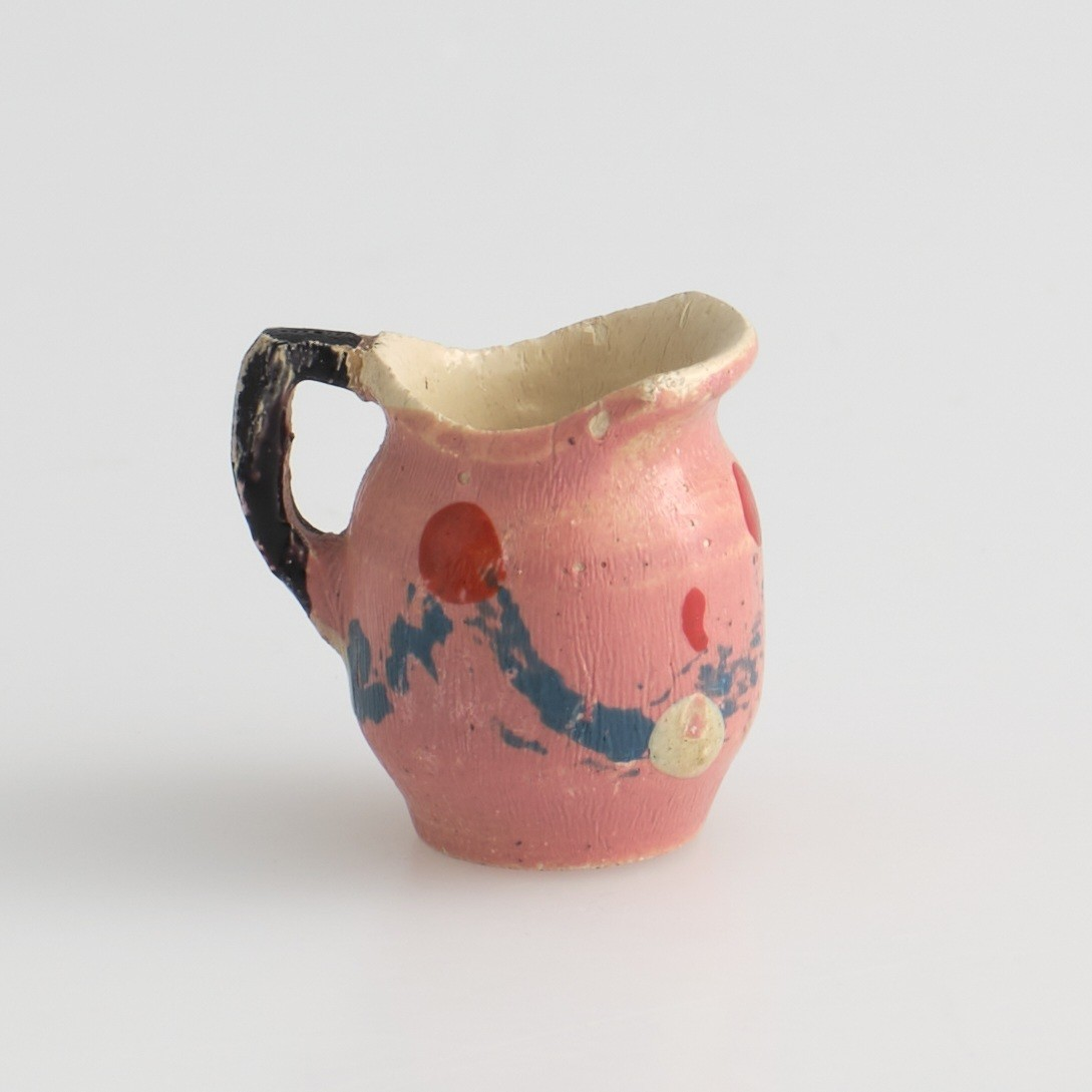 Serveringssett i miniatyr, lagd av tre.
Settet består av en tallerken, en kopp, en mugge og en krukke. Hele settet er malt i samme farger dekorert med likt mønster, utenom tallerkenen som ikke har malt mønster.
Tallerkenen har forhøyet kanter, og er malt rosa på innsiden, med hvite kanter og hvit underside.
Koppen er malt hvit på innsiden, og har en svartmalt hank. Hanken er øreformet og er festet fra kanten øverst til hvor koppen buer seg innover ved foten. Utsiden av koppen er malt rosa, og dekorert med blåe streker som danner trekanter med ujevne linjer rundt koppen. På toppen av trekantene er det malt en rød sirkel.
Krukken er sylinder-formet og snevres inn i mot toppen, før den vies skrått utover rundt åpningen. Krukken er malt hvit på innsiden, og rosa på utsiden, utsiden er også dekorert med samme mønster som koppen. Kanten rundt krukkens åpning er delvis brukket av.
Muggen er sylindret-formet, og snevres inn mot toppen, hvor kanten rundt åpningen forhøyes mot helletuten på den ene siden. På motsatt side av helletuten er det festet en svart-malt hank. Oversiden av hanken er rett, og bues ned mot bunnen av muggen. Muggen er malt og dekorert likt som koppen.