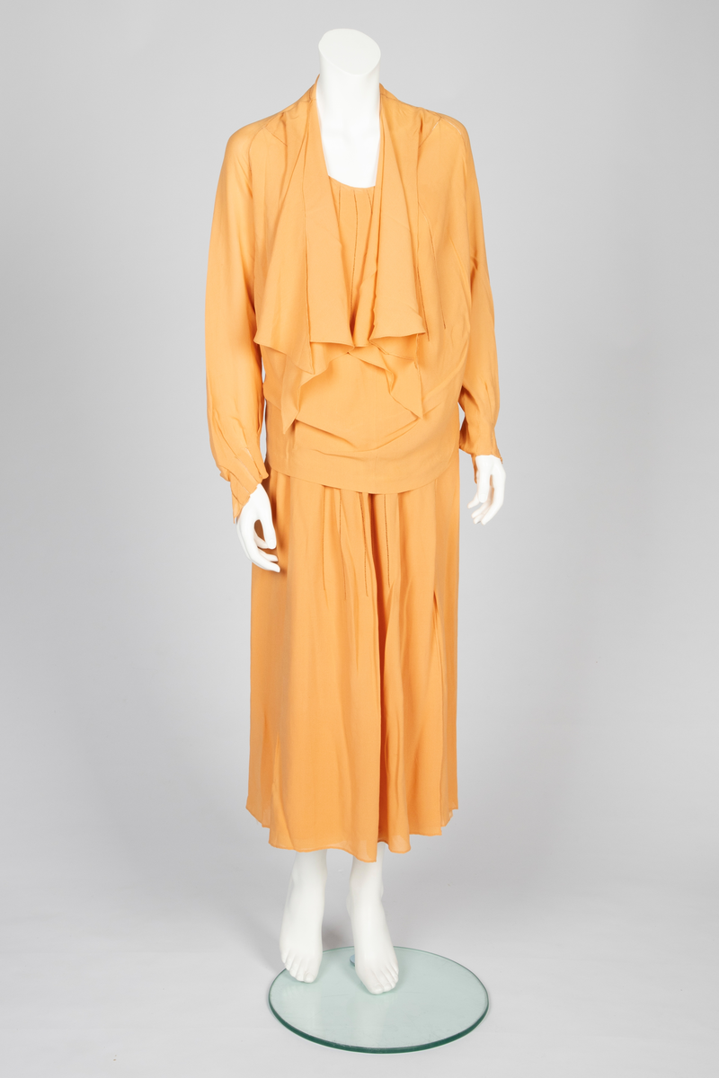 Todelt kjole bestående av et skjørt med påsydd underkjole og en løsthengende bluse. Skjørtet er legglang med rett fasong og er i hoftehøyde sydd til underkjole med stropper over skuldrene. Skjørtet har en rett bredde midt foran, inn mot denne er det lagt en fold på hver side. Herover er et løsthengende panel med sju striper dobbelt hullsøm, vertikalt og vifteformet, sydd på panelets øvre del. Blusen har søm midt foran og kile i hver side. Lange raglanermer med små folder og hullsøm ved håndleddene. Rundet halsringning foran, høy bak. Fra hals og skulderpartiet faller et løst, nærmest trekantet skjerf fremover på hver side. Hullsømstriper over brystet tilsvarende skjørtets.