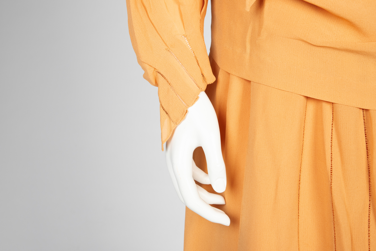 Todelt kjole bestående av et skjørt med påsydd underkjole og en løsthengende bluse. Skjørtet er legglang med rett fasong og er i hoftehøyde sydd til underkjole med stropper over skuldrene. Skjørtet har en rett bredde midt foran, inn mot denne er det lagt en fold på hver side. Herover er et løsthengende panel med sju striper dobbelt hullsøm, vertikalt og vifteformet, sydd på panelets øvre del. Blusen har søm midt foran og kile i hver side. Lange raglanermer med små folder og hullsøm ved håndleddene. Rundet halsringning foran, høy bak. Fra hals og skulderpartiet faller et løst, nærmest trekantet skjerf fremover på hver side. Hullsømstriper over brystet tilsvarende skjørtets.