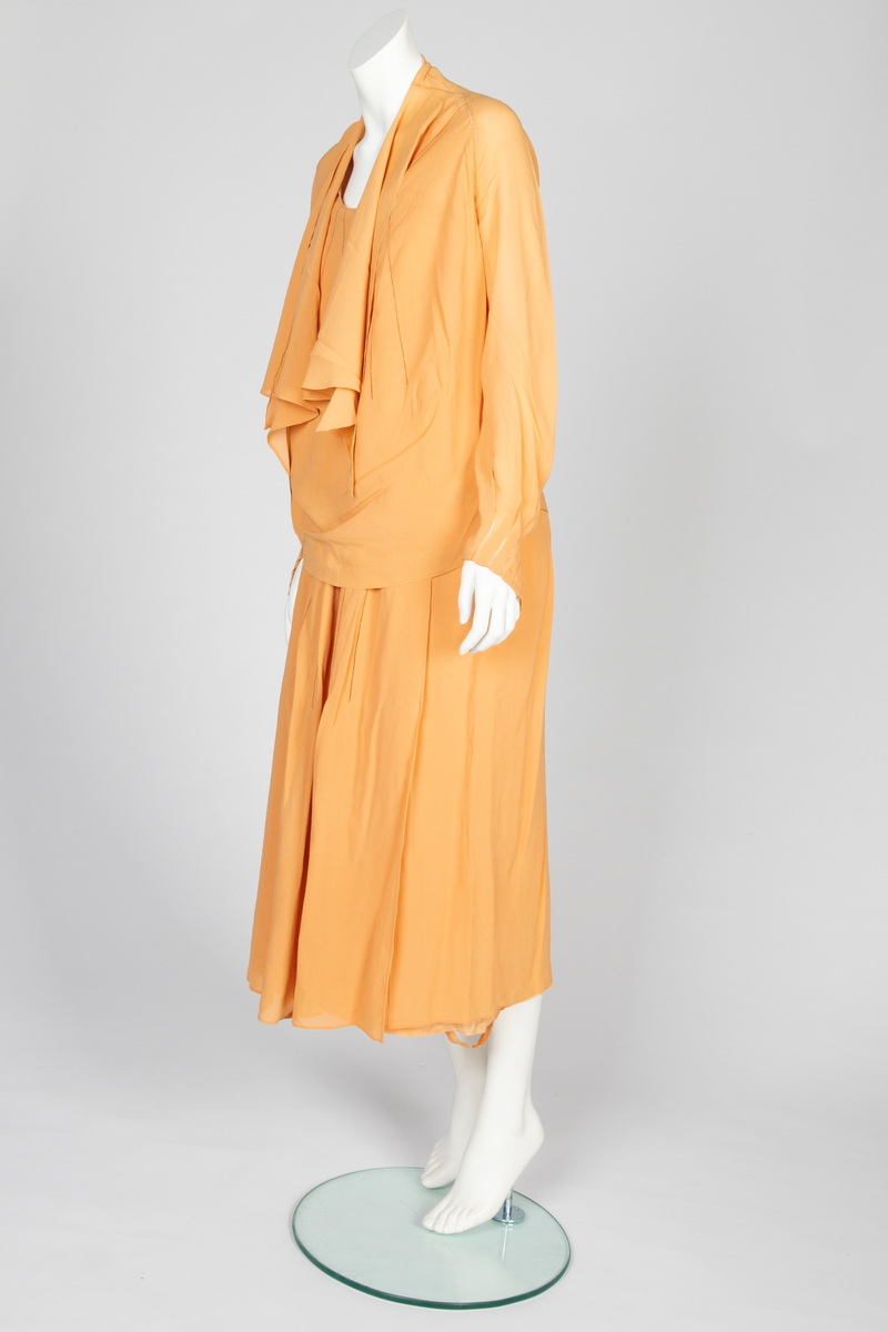 Todelt kjole bestående av et skjørt med påsydd underkjole og en løsthengende bluse. Skjørtet er legglang med rett fasong og er i hoftehøyde sydd til underkjole med stropper over skuldrene. Skjørtet har en rett bredde midt foran, inn mot denne er det lagt en fold på hver side. Herover er et løsthengende panel med sju striper dobbelt hullsøm, vertikalt og vifteformet, sydd på panelets øvre del. Blusen har søm midt foran og kile i hver side. Lange raglanermer med små folder og hullsøm ved håndleddene. Rundet halsringning foran, høy bak. Fra hals og skulderpartiet faller et løst, nærmest trekantet skjerf fremover på hver side. Hullsømstriper over brystet tilsvarende skjørtets.