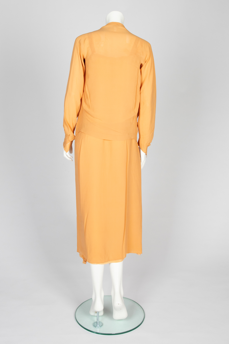 Todelt kjole bestående av et skjørt med påsydd underkjole og en løsthengende bluse. Skjørtet er legglang med rett fasong og er i hoftehøyde sydd til underkjole med stropper over skuldrene. Skjørtet har en rett bredde midt foran, inn mot denne er det lagt en fold på hver side. Herover er et løsthengende panel med sju striper dobbelt hullsøm, vertikalt og vifteformet, sydd på panelets øvre del. Blusen har søm midt foran og kile i hver side. Lange raglanermer med små folder og hullsøm ved håndleddene. Rundet halsringning foran, høy bak. Fra hals og skulderpartiet faller et løst, nærmest trekantet skjerf fremover på hver side. Hullsømstriper over brystet tilsvarende skjørtets.