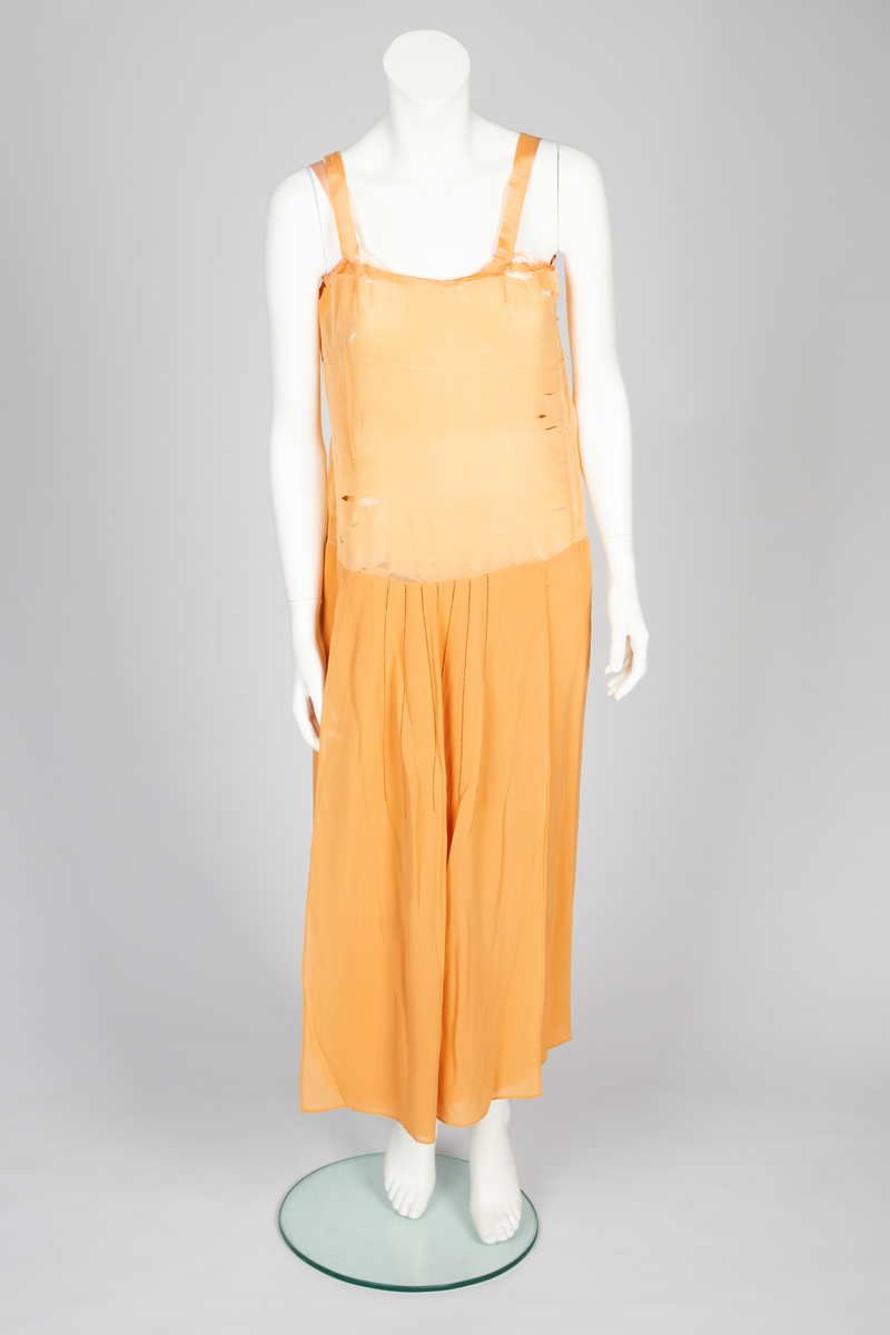 Todelt kjole bestående av et skjørt med påsydd underkjole og en løsthengende bluse. Skjørtet er legglang med rett fasong og er i hoftehøyde sydd til underkjole med stropper over skuldrene. Skjørtet har en rett bredde midt foran, inn mot denne er det lagt en fold på hver side. Herover er et løsthengende panel med sju striper dobbelt hullsøm, vertikalt og vifteformet, sydd på panelets øvre del. Blusen har søm midt foran og kile i hver side. Lange raglanermer med små folder og hullsøm ved håndleddene. Rundet halsringning foran, høy bak. Fra hals og skulderpartiet faller et løst, nærmest trekantet skjerf fremover på hver side. Hullsømstriper over brystet tilsvarende skjørtets.