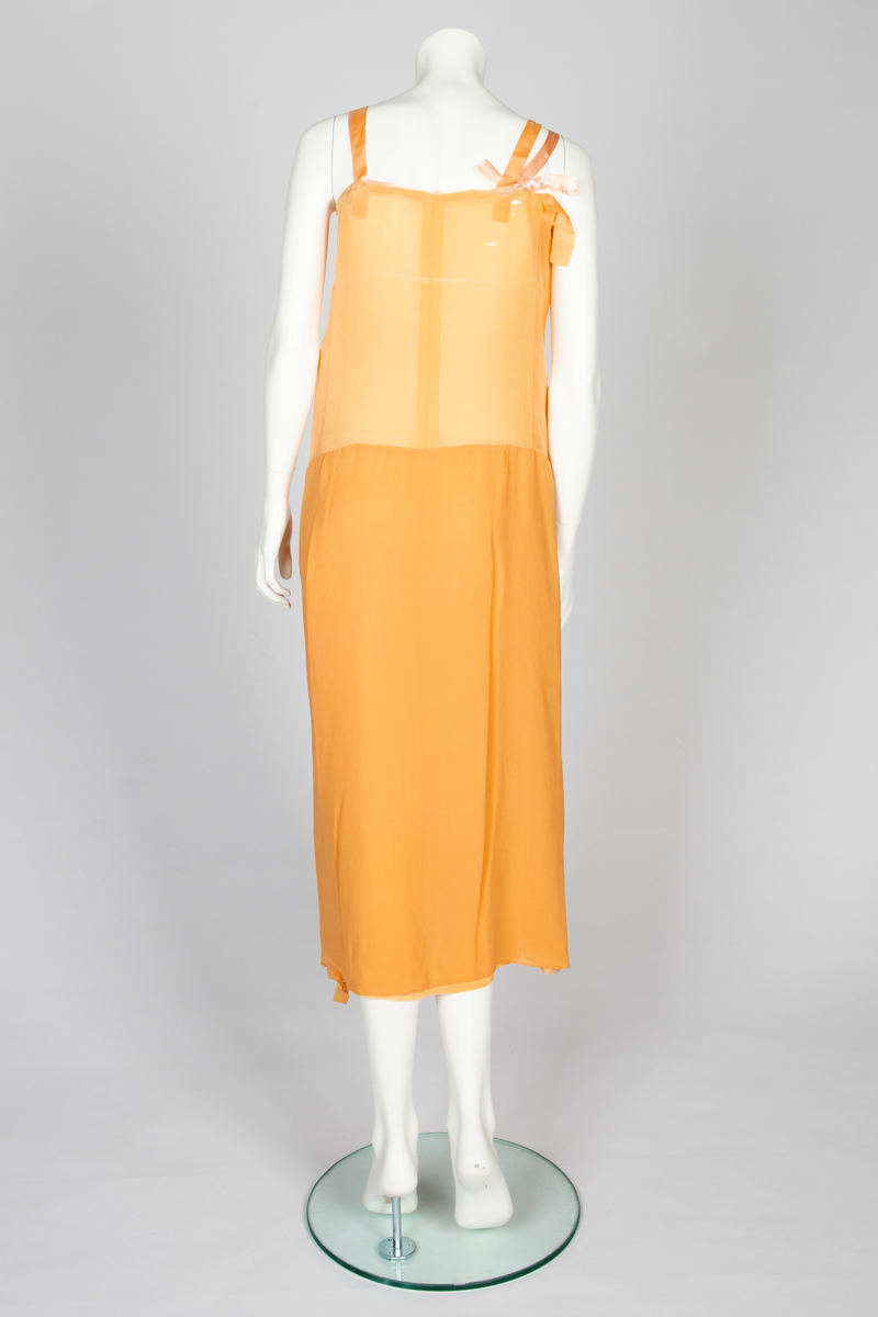 Todelt kjole bestående av et skjørt med påsydd underkjole og en løsthengende bluse. Skjørtet er legglang med rett fasong og er i hoftehøyde sydd til underkjole med stropper over skuldrene. Skjørtet har en rett bredde midt foran, inn mot denne er det lagt en fold på hver side. Herover er et løsthengende panel med sju striper dobbelt hullsøm, vertikalt og vifteformet, sydd på panelets øvre del. Blusen har søm midt foran og kile i hver side. Lange raglanermer med små folder og hullsøm ved håndleddene. Rundet halsringning foran, høy bak. Fra hals og skulderpartiet faller et løst, nærmest trekantet skjerf fremover på hver side. Hullsømstriper over brystet tilsvarende skjørtets.