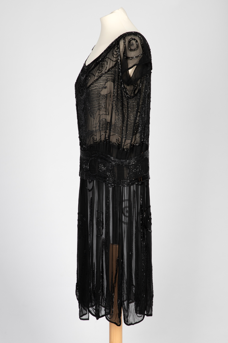 Knekort perlekjole i svart, transparent slikechiffon med rund hals og korte ermer. Rikt, svart perle- og paljettbroderi med motiv av steilende hester opp av bølger.