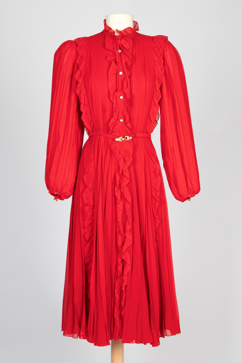 Kort, plissert chiffonkjole med lange, posete ermer og vertikale rysjer over kjolens for- og bakstykket. Knappestolpe med dekorative strassknapper foran og oppstående rysjekant langs halspningen. Smalt belte i kjolestoffet med gullfarget beltespenne dekorert med strasstener.