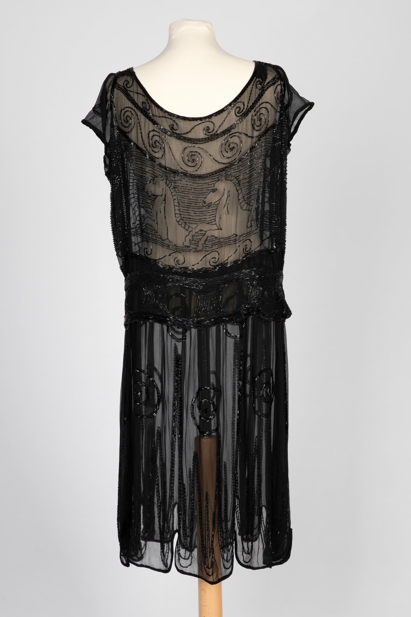 Knekort perlekjole i svart, transparent slikechiffon med rund hals og korte ermer. Rikt, svart perle- og paljettbroderi med motiv av steilende hester opp av bølger.