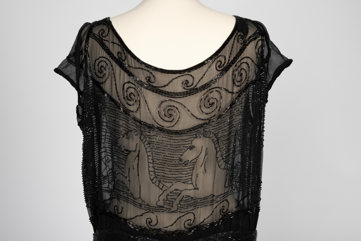 Knekort perlekjole i svart, transparent slikechiffon med rund hals og korte ermer. Rikt, svart perle- og paljettbroderi med motiv av steilende hester opp av bølger.