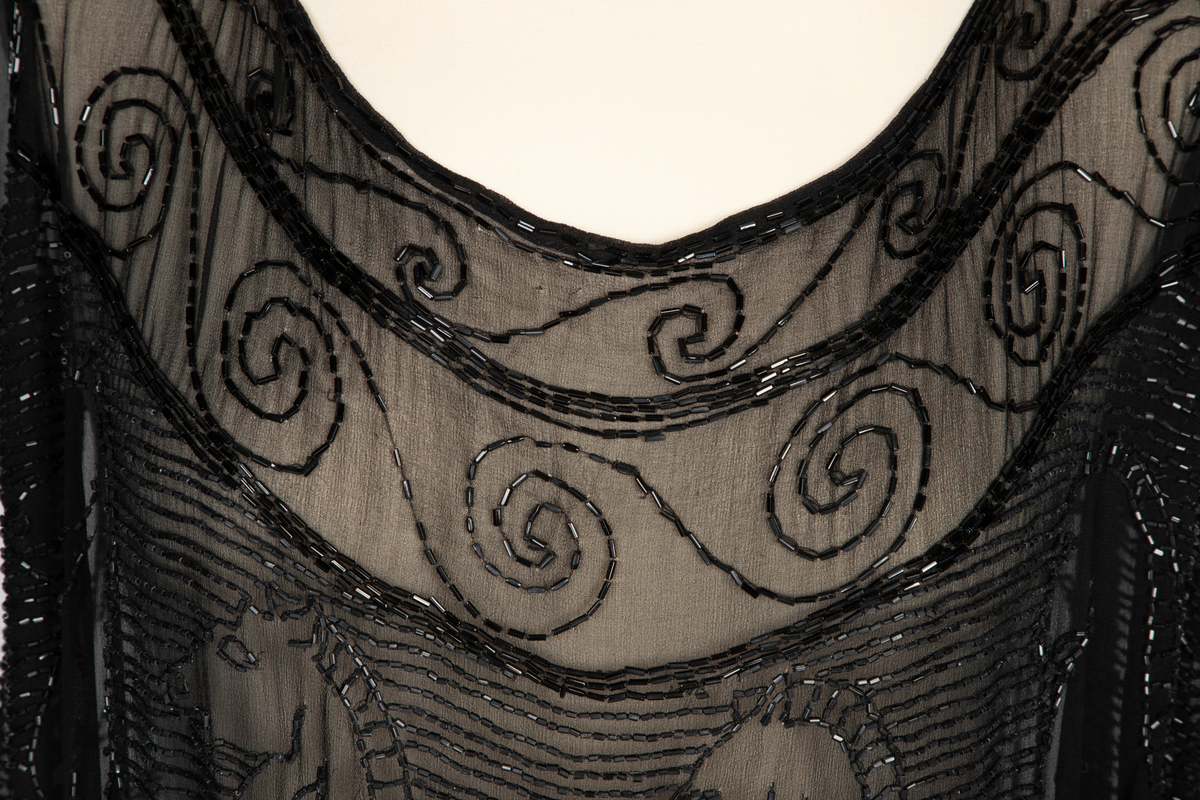 Knekort perlekjole i svart, transparent slikechiffon med rund hals og korte ermer. Rikt, svart perle- og paljettbroderi med motiv av steilende hester opp av bølger.