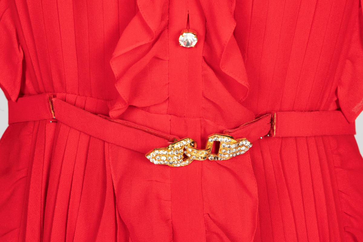 Kort, plissert chiffonkjole med lange, posete ermer og vertikale rysjer over kjolens for- og bakstykket. Knappestolpe med dekorative strassknapper foran og oppstående rysjekant langs halspningen. Smalt belte i kjolestoffet med gullfarget beltespenne dekorert med strasstener.