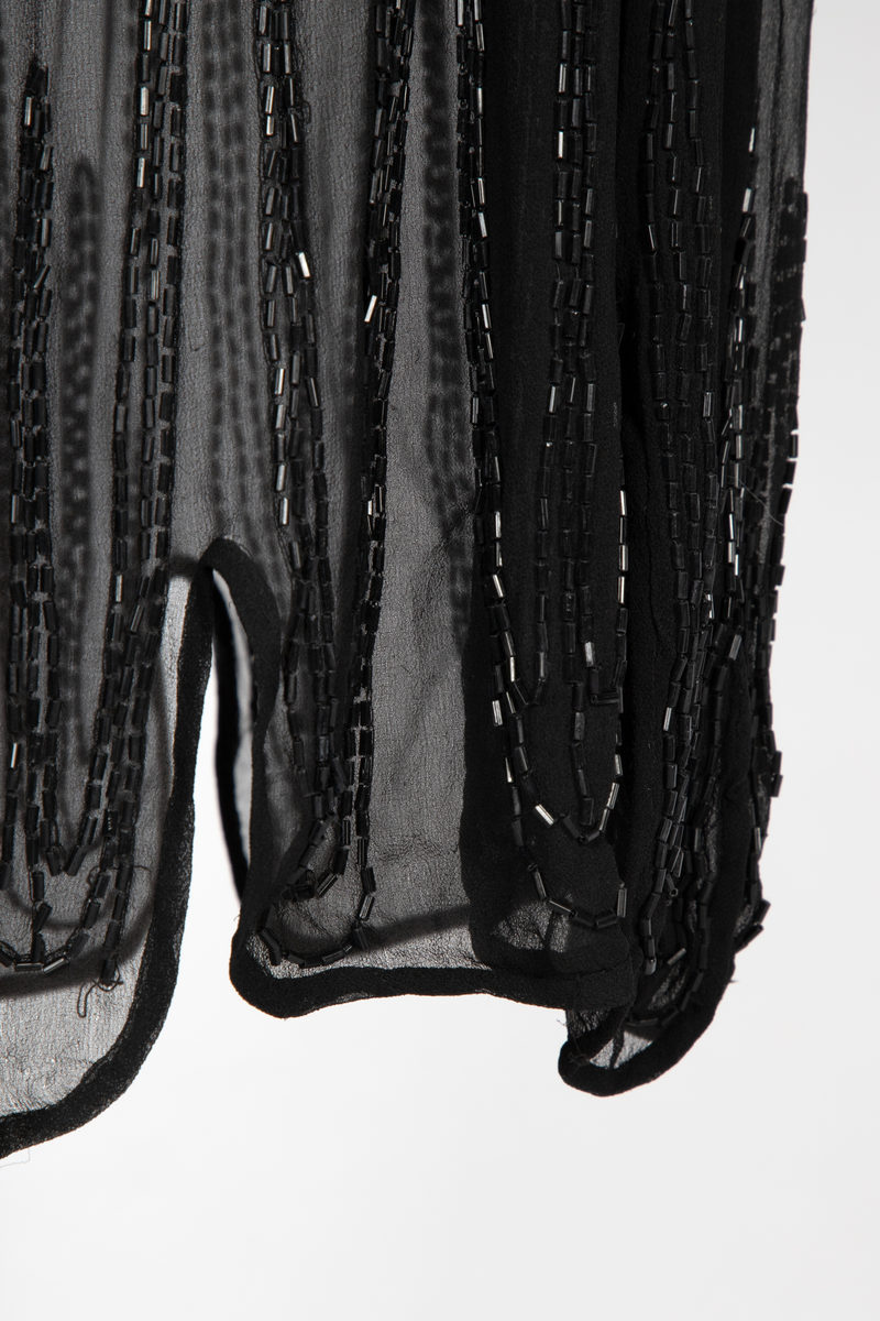 Knekort perlekjole i svart, transparent slikechiffon med rund hals og korte ermer. Rikt, svart perle- og paljettbroderi med motiv av steilende hester opp av bølger.