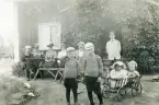 Ett sällskap har slagit sig ner framför en stuga. 
Längst t. v. syns kronolänsman Albert Bergstedt. I vit blus hans fru Elisabeth Bergstedt. 
Mellan de två pojkarna i randiga tröjor syns stadsfiskal Filip Rehnström. Flickan i kärran är Margareta Bergstedt.