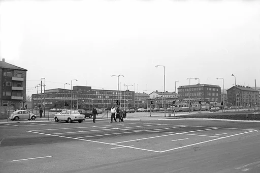 Fotografiet visar en vy som visar stadshuset och på avstånd platsen där Mölndals stadsbibliotek och Folkets hus ligger samlade.
Människor går på trottoarer och personbilar kör i riktningar både till och från Göteborg.

Två sammankopplade spårvagnar är på väg in mot Mölndalsbro.

Det kan vara svårt att se, men om man tittar nära, till vänster om stadshuset, så syns byggnaden för Mölndals taxistation.