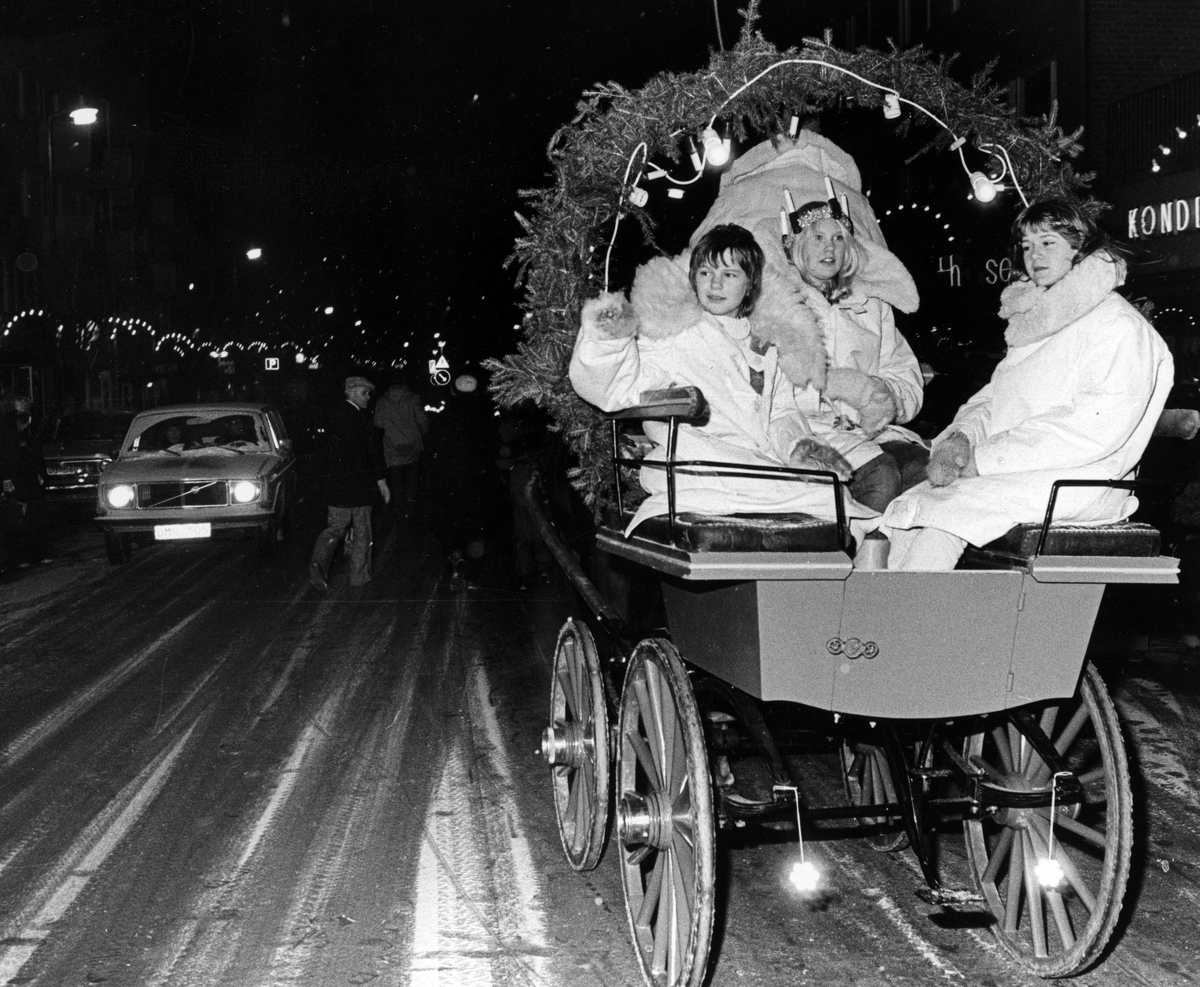 Nynäshamns lucia. Beriden luciakortege som går på Centralgatan i Nynäshamns centrum. Lucia sitter i en vagn, tillsammans med henne sitter två ungdomar. Alla klädda i vita ytterrockar med skinnkrage. 