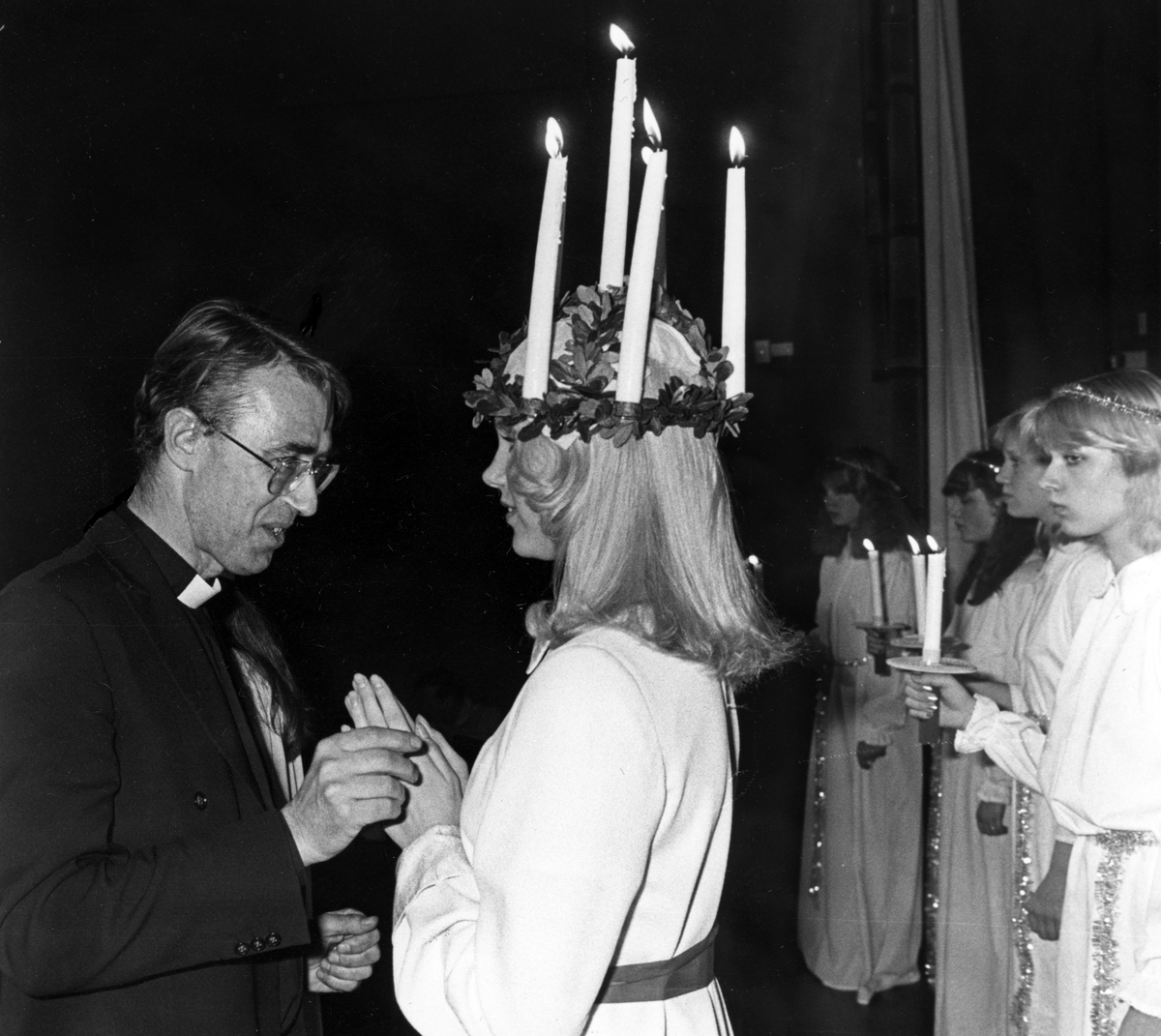 Kyrkoherden Sten Andræ kröner Nynäshamns lucia, en ljushårig ung kvinna med en luciakrona med levande ljus. Bakom henne syns några tärnor som håller i ljus. 