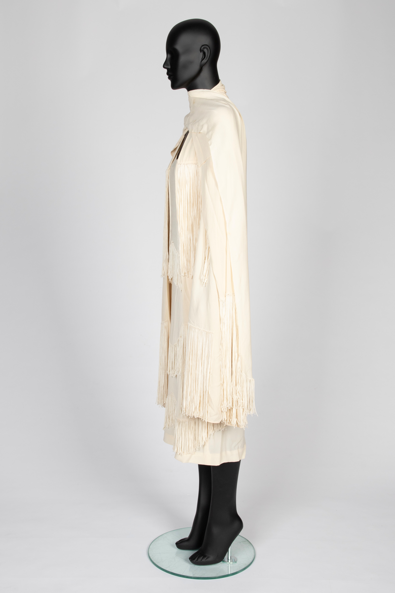 Ensemble i elfenbensfarget silkekrepp (crêpe georgette) med eggeskallhvite snorfrynser. Ensemblen består av tre deler; kjole, cape og skjerf. Kjolen er ermeløs, har rett fasong og vid utringning. Skjørtet er festet til et underliv, med en løsthengende bluse over, bare festet til underlivet bak. Blusen er tunget og har frynser nederst. Skjørtet har to løsthengende overskjørt eller kapper i forskjellig lengde, begge festet i livsømmen og med tunget avslutning og frynser. Capen er vid og i samme stoff, fôret med et bredt, kragelignende skjerf, som faller frem over skuldrene i lange, brede skjerfender med lange frynser. I ryggen to lange frynserader. Den nedre faller helt til capens nederkant. Langt skjerf med rundede avslutninger og frynser.