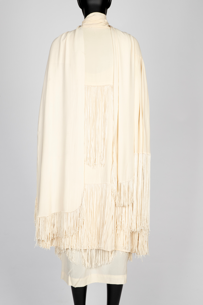 Ensemble i elfenbensfarget silkekrepp (crêpe georgette) med eggeskallhvite snorfrynser. Ensemblen består av tre deler; kjole, cape og skjerf. Kjolen er ermeløs, har rett fasong og vid utringning. Skjørtet er festet til et underliv, med en løsthengende bluse over, bare festet til underlivet bak. Blusen er tunget og har frynser nederst. Skjørtet har to løsthengende overskjørt eller kapper i forskjellig lengde, begge festet i livsømmen og med tunget avslutning og frynser. Capen er vid og i samme stoff, fôret med et bredt, kragelignende skjerf, som faller frem over skuldrene i lange, brede skjerfender med lange frynser. I ryggen to lange frynserader. Den nedre faller helt til capens nederkant. Langt skjerf med rundede avslutninger og frynser.