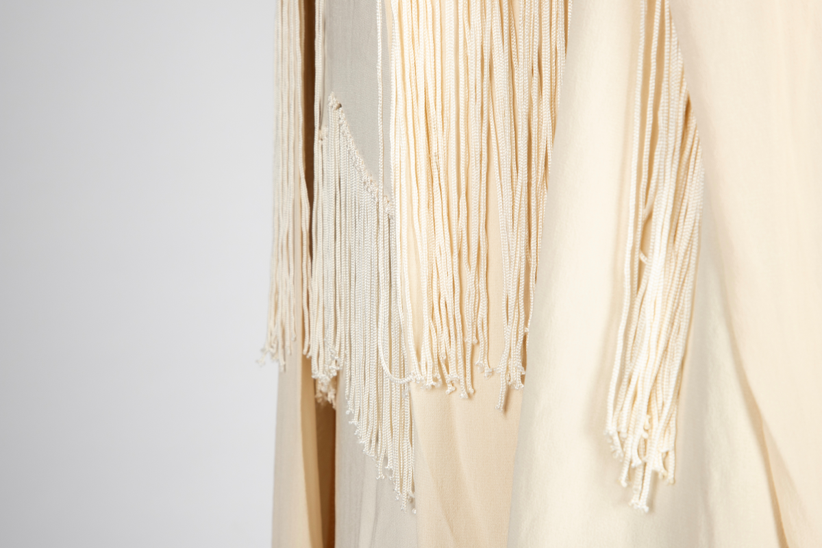 Ensemble i elfenbensfarget silkekrepp (crêpe georgette) med eggeskallhvite snorfrynser. Ensemblen består av tre deler; kjole, cape og skjerf. Kjolen er ermeløs, har rett fasong og vid utringning. Skjørtet er festet til et underliv, med en løsthengende bluse over, bare festet til underlivet bak. Blusen er tunget og har frynser nederst. Skjørtet har to løsthengende overskjørt eller kapper i forskjellig lengde, begge festet i livsømmen og med tunget avslutning og frynser. Capen er vid og i samme stoff, fôret med et bredt, kragelignende skjerf, som faller frem over skuldrene i lange, brede skjerfender med lange frynser. I ryggen to lange frynserader. Den nedre faller helt til capens nederkant. Langt skjerf med rundede avslutninger og frynser.