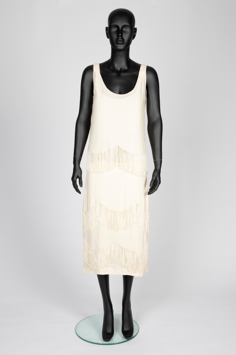 Ensemble i elfenbensfarget silkekrepp (crêpe georgette) med eggeskallhvite snorfrynser. Ensemblen består av tre deler; kjole, cape og skjerf. Kjolen er ermeløs, har rett fasong og vid utringning. Skjørtet er festet til et underliv, med en løsthengende bluse over, bare festet til underlivet bak. Blusen er tunget og har frynser nederst. Skjørtet har to løsthengende overskjørt eller kapper i forskjellig lengde, begge festet i livsømmen og med tunget avslutning og frynser. Capen er vid og i samme stoff, fôret med et bredt, kragelignende skjerf, som faller frem over skuldrene i lange, brede skjerfender med lange frynser. I ryggen to lange frynserader. Den nedre faller helt til capens nederkant. Langt skjerf med rundede avslutninger og frynser.